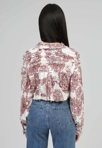 Femme aux longs cheveux noirs portant une veste courte à motifs paisley blanche et bordeaux ainsi que des jeans en denim bleu, de dos sur un fond gris.