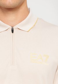 Bežna polo majica z zadrgo na vratu, s svetlo rumenim poudarkom na ovratniku in rumenim vezenim logotipom na prsih.