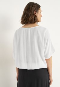 Witte, lichte blouse met een textuurafwerking en losse, korte mouwen; ronde halslijn en een cropped achterkant. Gedomineerd met een zwarte rok.