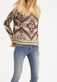 Beige stickad tröja med bruna geometriska mönster, rund hals, långa ärmar, kombinerad med mid-blue jeans. Mjuk struktur och avslappnad passform.