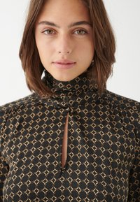 Schwarz gemustertes Kleid mit hohem Kragen, Schlüsselloch-Ausschnitt und goldenen geometrischen Designs. Glatte Textur und figurbetonte Silhouette.