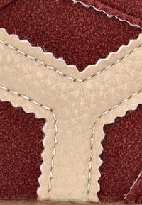 Zapatilla de cuero con una parte superior en color burdeos y acentos en color crema. Presenta costuras en zigzag y superficies texturizadas, resaltando un diseño geométrico.