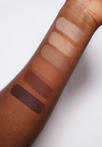 Stalen van zes foundationshuidskleuren op een bruine arm, variërend van lichtbeige tot diep chocolade. Gladde textuur met een matte afwerking.