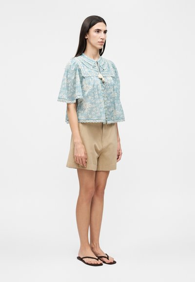 Νέα γυναίκα που φοράει ένα ανοιχτό μπλε φλοράλ πουκάμισο με δαντελένιες λεπτομέρειες, μπεζ σορτς και μαύρα σαγιονάρια, στέκεται μπροστά σε λευκό φόντο.