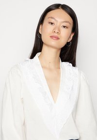 Blusa branca com decote em V e acabamento em renda decorativa. Mangas largas e um tecido texturizado caracterizado por um material suave e respirável.