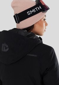 Vrouw draagt een lichtroze gebreide muts, skibril en een zwarte capuchonjas met zichtbare logo's van "Smith" en "Fundango".