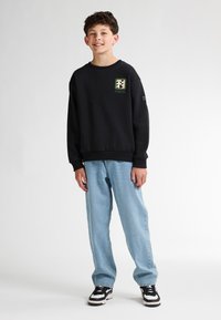 Sweatshirt noir avec design de logo sur la poitrine, associé à des jeans baggy bleu clair et des baskets noir et blanc. Fond uni.