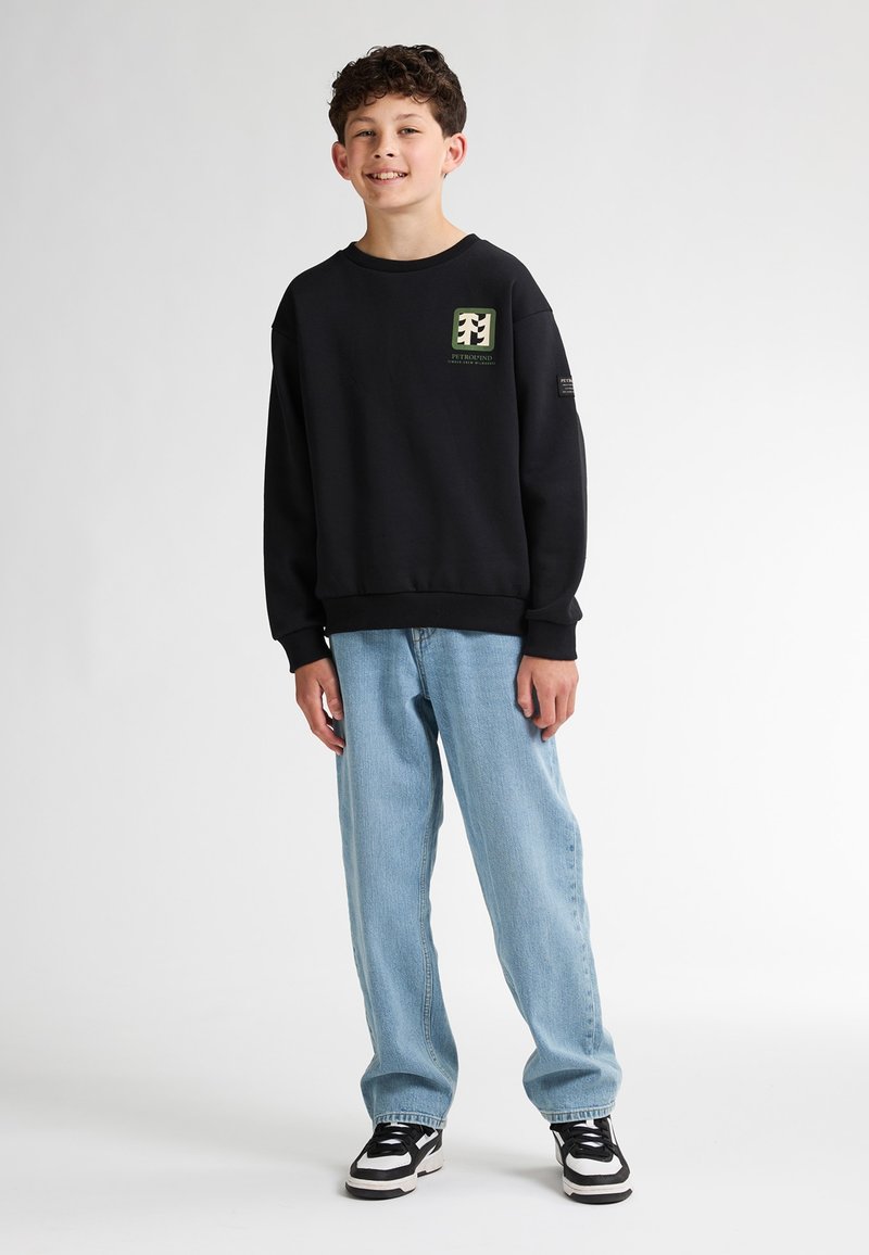 Sweatshirt noir avec design de logo sur la poitrine, associé à des jeans baggy bleu clair et des baskets noir et blanc. Fond uni.