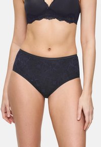 Femeie îmbrăcată într-un sutien din dantelă neagră și chiloți din dantelă cu talie înaltă asortați, arătând secțiunea medie și partea superioară a coapselor pe un fundal simplu.