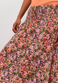 Jupe longue fleurie avec une taille smockée, des motifs colorés en rose, orange et vert, en tissu léger et au design fluide.