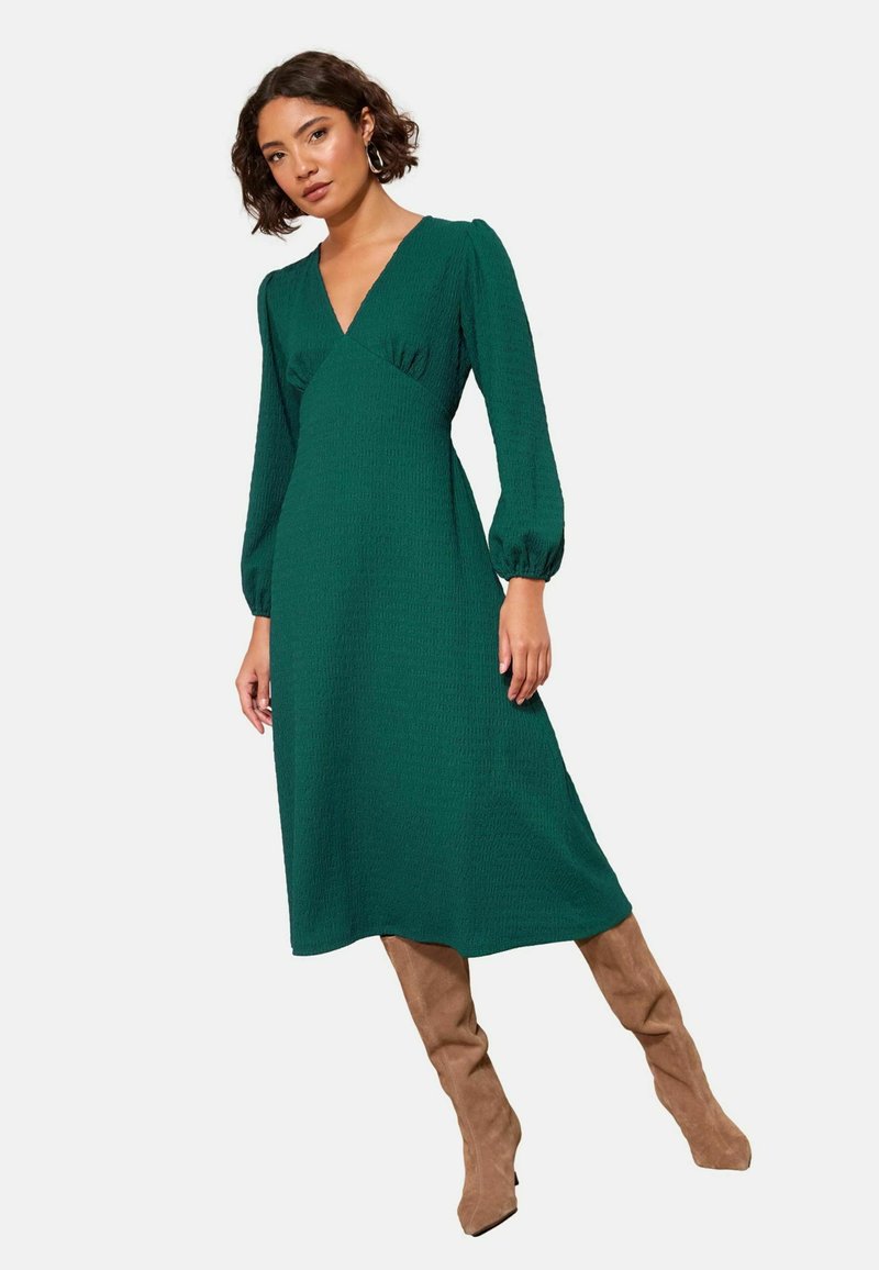 Rochie verde, texturată, de lungime midi, cu decolteu în V și mâneci bufante, asortată cu cizme bej până la genunchi. Fără modele sau accesorii vizibile.