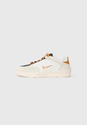 Baskets basses Nike blanches et beiges avec un swoosh orange, des lacets orange et noirs, et une semelle beige clair sur un fond gris uni.