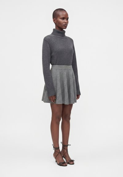 Femme debout portant un pull à col roulé gris, une jupe plissée grise et des sandales noires à talons hauts avec des brides à la cheville, sur un fond uni.
