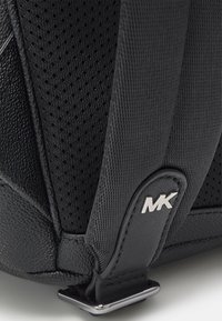 Sac noir en cuir texturé et en mesh, avec un détail de logo MK argenté sur une sangle et des éléments de design angulaires.