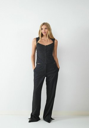 Młoda kobieta stojąca z rękami w kieszeniach, ubrana w czarny bezrękawnik w drobną prążek i pasujące luźne spodnie o szerokich nogawkach na jednolitym białym tle.