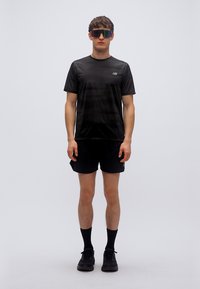 Μαύρο αθλητικό t-shirt με κοντά μανίκια, λογότυπο στο στήθος, συνδυασμένο με μαύρο σόρτς και αθλητικά μαύρα παπούτσια. Φοράει καθρεφτιστά γυαλιά ηλίου.