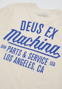 Beige T-shirt med iøjnefaldende blå tekst, der læser "DEUS EX Machina DXM PARTS & SERVICE USA LOS ANGELES, CA" i forskellige skrifttyper.