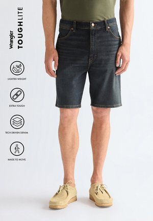 Man met donkere denimshorts en beige casual schoenen, staand met ontspannen handen langs het lichaam tegen een effen lichte achtergrond.