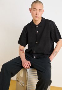 Black short-sleeve knitted polo shirt with a textured pattern, plastic buttons, and relaxed fit, paired with black trousers.