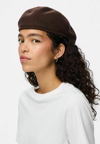 Boina de fieltro marrón con forma redondeada, combinada con una camiseta blanca. Presenta una tela texturizada y un acabado suave.