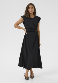 Crna haljina bez rukava s bočnim prorezom, s detaljem kopče u struku. Glatka tkanina s opuštenim krojem. Kombinirana s prugastim potpeticama.