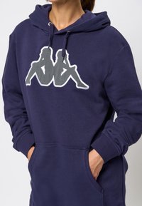 Personne portant un sweat à capuche bleu marine avec une poche avant et un logo gris et blanc représentant deux silhouettes assises dos à dos sur la poitrine.