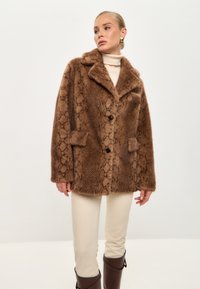 Brauner Kunstpelzmantel mit Schlangenmuster, mit von hinten offenen Kragen, zwei vorderen Taschen und zwei dunklen Knöpfen. Kombiniert mit cremefarbenen Hosen.