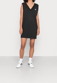 Svart ärmlös klänning med V-ringning, tillverkad av bomullsblandat material. Har en broderad logotyp på bröstet. Enkel och rak form.