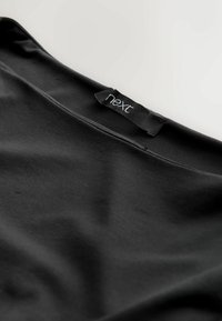 Top in tessuto nero con una texture liscia, dotato di un'etichetta con la scritta "next" sul colletto. Design semplice senza ulteriori motivi o dettagli.
