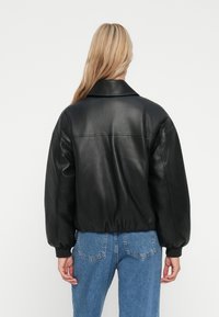 Frau mit langen blonden Haaren, die eine schwarze Lederjacke mit elastischem Saum und Bluejeans trägt, mit dem Rücken zu einer weißen Wand.