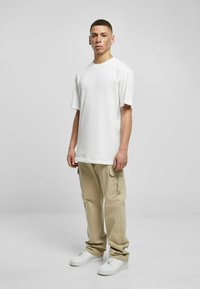 Camiseta blanca de manga corta combinada con pantalones cargo beige claros y zapatillas blancas, de pie frente a un fondo simple.