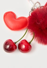 Μπρελόκ καρδιά από κόκκινο δέρμα με δύο λαμπερά charms κεράσι και ένα κόκκινο διακοσμητικό φούντα από συνθετική γούνα, προσαρτημένο σε μεταλλικό κούμπωμα.