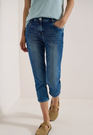 Personne portant un jean bleu court et des chaussures à boucle marron clair, une main dans la poche, debout devant un fond clair uni.