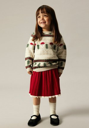 Maglione a maglia di colore crema con motivi a tema natalizio, gonna rossa a campana, calze bianche fino al ginocchio e scarpe Mary Jane nere. Sfondo pulito.