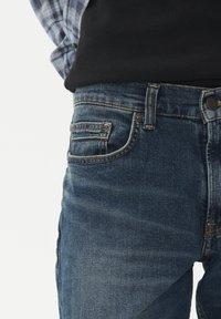 Artimiausias vaizdas asmens, kuris dėvi mėlynus džinsus su priekinėmis kišenėmis ir diržo kilpa, derinant juodą viršų ir mėlyną languotą marškinį.