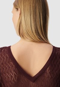 Bordeaux V-hals trui met een ruitpatroon en gebreide textuur. Haar valt over de schouder, met een gladde huid zichtbaar bij de halslijn.