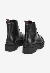 Botas de cuero negro hasta el tobillo con suela gruesa, diseño con cordones y una lengüeta a rayas contrastantes en el talón. Textura lisa en toda la bota.