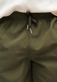Gros plan sur la taille d’un pantalon vert olive à cordon avec des liens noirs et des embouts métalliques argentés, associé à un haut en maille blanc.