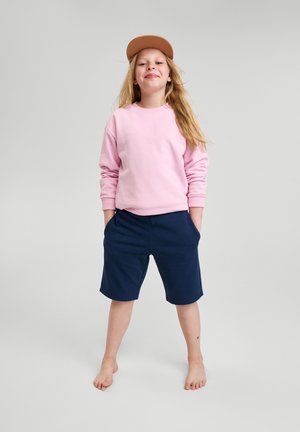 Jasnoróżowy bawełniany sweatshirt, granatowe szorty i beżowa czapka. Prosty design, luźny krój. Stojąca bosa postawa na szarym tle.