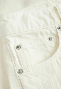 Tessuto in denim bianco con una tasca posteriore dotata di rivetti metallici argentati, una superficie texturizzata e cuciture parallele lungo i bordi.