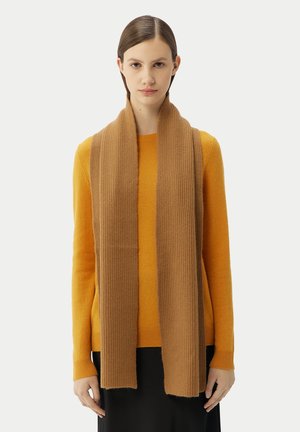 Frau mit glattem braunem Haar trägt einen langen, gerippten Schals in Kamelhaarfarbe über einem senffarbenen Pullover und einem schwarzen Rock und steht vor einem schlichten Hintergrund.