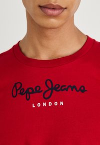 Rød bomulds T-shirt med et sort skrifttype-logo "Pepe Jeans" over ordet "LONDON" i hvid. Rund halsudskæring, korte ærmer.