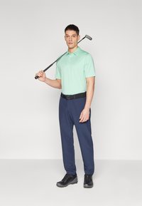 Lys grønn polo skjorte, marine bukser og svarte sko. Poloen har en klassisk krage; buksene har rett snitt. En golfklubb holdes.