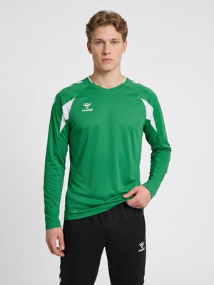 Młody mężczyzna w zielonej sportowej koszulce z długim rękawem i czarnych spodniach, stoi z neutralnym wyrazem twarzy na jednolitym tle.