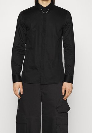 Personne portant une chemise noire cintrée à manches longues avec une chaîne décorative au col et un pantalon cargo noir avec poches latérales, debout sur un fond neutre.