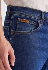 Gros plan d’une personne portant un jean en denim bleu foncé avec des coutures visibles et un petit logo Wrangler en cuir sur la poche avant.