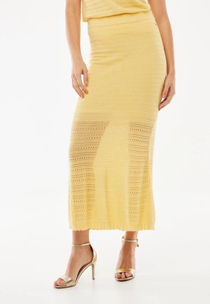 Kobieta ubrana w żółtą dzianinową midisukienkę w parze ze złotymi sandałami na wysokim obcasie, stojąca na tle jednolitego tła.