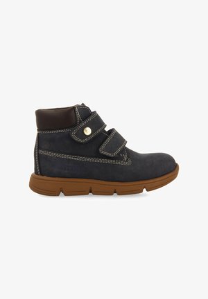 Bottines en suède bleu marine avec deux bandes Velcro, semelle en caoutchouc beige, coutures blanches contrastantes et un col en cuir marron lisse.