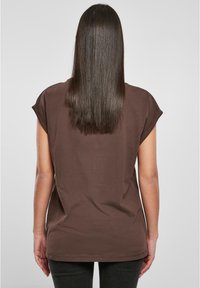 Frau mit glattem, dunklem Haar, die ein kurzärmliges braunes Shirt trägt und mit dem Rücken vor einem schlichten hellgrauen Hintergrund steht.