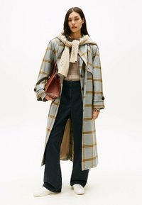 Abrigo largo de cuadros en azul y amarillo, combinado con un suéter beige de punto trenzado y pantalones anchos oscuros. Bolso marrón y zapatillas blancas.